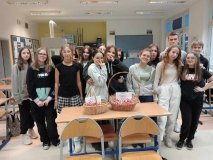 Wielu uczestników Koła zgromadzonych w jednym miejscu sali, wszyscy uśmiechnięci i przyjaźnie nastawieni, przed nimi na ławce znajdują się wiklinowe koszyczki, całe wpełnione baiło-czerwonymi kotylionami