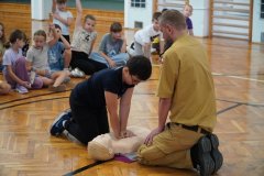 Chłopiec, instruowany przez strażaka, wykonujący resuscytację na fantomie. W oddali grupa dzieci.