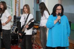 Kobieta z mikrofonem przemawia podczas szkolnego występu. Obok znajdują się Uczniowie, z gitarą i perkusją, przygotowani do koncertu.