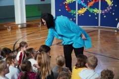 Kobieta pochyla się z mikrofonem nad grupą Dzieci siedzących na podłodze podczas szkolnego wydarzenia. W tle znajduje się dekoracja z symbolem nieskończoności.