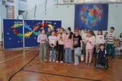 Grupa Uczniów występuje na sali gimnastycznej, śpiewając do mikrofonu. W tle widnieje dekoracja z symbolem nieskończoności i hasłem dotyczącym różnorodności ludzi.