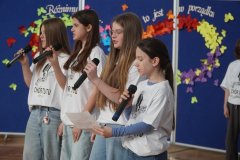 Cztery Dziewczynki stoją obok siebie i śpiewają do mikrofonów. Jedna z nich trzyma kartkę z tekstem. Ubrane są w białe koszulki z nadrukiem nazwy chóru, a w tle widać kolorową dekorację z motylami i napisem na niebieskiej tablicy.