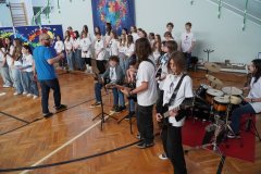 W sali gimnastycznej  znajdują się Uczniowie  tworzący chór i zespół muzyczny – część z nich śpiewa, a inni grają na gitarach i perkusji. Nauczyciel stoi przed nimi i prowadzi występ, a w tle widać dekorację z kolorowym motywem puzzli.