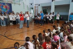 W sali gimnastycznej odbywa się występ szkolny – na środku stoją Uczniowie z mikrofonami, a za nimi chór i zespół muzyczny z gitarami. Przed sceną siedzi grupa Dzieci, która ogląda występ.