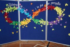 Niebieska tablica dekoracyjna ozdobiona kolorowymi papierowymi motylami tworzącymi tęczowe wzory. Na górze widnieje napis: „Różnimy się… i to jest w porządku”.