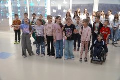 Grupa Dzieci śpiewa. Część z nich trzyma mikrofony. Wśród Uczestników jest także Chłopiec na wózku inwalidzkim. W tle widać innych Uczniów oraz dekorację z niebieskimi elementami zawieszonymi przy oknach.