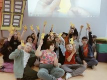 Gupa uczniów w bibliotece, siedzią „po turecku” na podłodze, w rękach trzymają żółte figurki origami – koty.