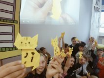 Grupa uczniów w bibliotece w wyciągniętych rękach prezentuje wykonane przez siebie żółte figurki origami – koty.