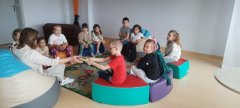 Grupa uczniów siedzi w kręgu wokół nauczycielki bibliotekarki, która czyta dzieciom książkę.