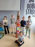 Grupa uczennic prezentuje zbudowaną z książek wieżę, która jest od nich wyższa „o głowę”, jedna uczennica siedzi z książką w ręku na podłodze.