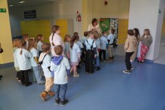 Grupa Dzieci w niebieskich koszulkach stoi na korytarzu szkolnym. Nauczyciel pokazuje Im szkołę, a w szczególności zwraca uwagę na kolorowe szafki, w których Uczniowie klas starszych pozostawiają swoje materiały szkolne.