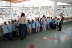 Grupa Dzieci w niebieskich koszulkach stoi w szeregu na I piętrze korytarza szkolnego i obserwuje zajęcia odbywające się na auli. Obok Dzieci stoją trzy Nauczycielki. Jedna z nich opowiada o zajęciach klas starszych.