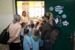 Grupa Dzieci w niebieskich koszulkach stoi w drzwiach klasy, w której odbędą się zajęcia. Zielone drzwi ozdobione są napisami-  „magiczne słowa”, „proszę” , „dziękuję” i „do widzenia”. Nauczycielka zaprasza Dzieci do sali.