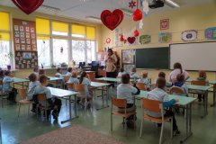 Dzieci siedzą przy stolikach klasowych odwróceni w stronę tablicy szkolnej i obserwują Nauczyciela, który przedstawia im Ucznia klasy III, który będzie pomagał im podczas zajęć. Nad głowami Dzieci umieszczona jest czerwona dekoracja w postaci serduszek i baloników.