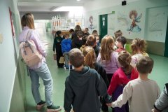Dzieci, ustawione parami, przechodzą korytarzem na piętrze i zmierzają w kierunku łącznika, aby zwiedzić pozostałą część szkoły. Po lewej stronie grupy znajduje się Nauczyciel.