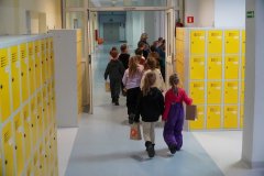 Grupa Dzieci znajduje się na korytarzu szkolnym, obok kolorowych szafek Uczniów klas starszych, i obserwuje zajęcia wychowania fizycznego odbywające się w sali gimnastycznej. Dziewczynka w białych spodniach i czarnej bluzie ma w ręku papierową torebeczkę z wykonaną pracą – kolorowym ślimakiem.