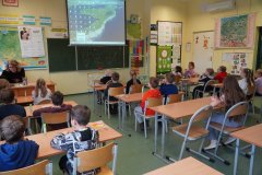 Dzieci siedzą w ławkach dwuosobowych i obserwują wyświetlaną przez nauczyciela prezentację. W tle widać kolorowe materiały dydaktyczne – mapa, tablice demonstracyjne.