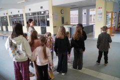 Grupa Dzieci stoi w szatni głównej i uważnie słucha wypowiedzi Nauczycielki, która opowiada o nauce w szkole.