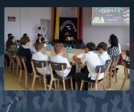 Zdjęcie przedstawia grupę uczniów siedzących wokół dużego, okrągłego stołu.                Na stole znajdują się napoje, przekąski oraz karteczki z imionami. W tle widoczny jest ekran z grafiką o szkolnej debacie w ramach programu „Szkoła Dobrego Słowa”.
