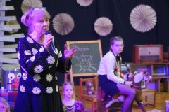 Nauczycielka przemawia do mikrofonu podczas szkolnego wydarzenia. W tle widać Dzieci biorące udział w przedstawieniu – jedno z nich siedzi na drewnianym koniku. Scena jest udekorowana papierowymi rozetami, zabawkami oraz tablicą z napisem „Dzień dobry”.