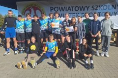 Na tle białego banneru szkolnego związku sportowego stoi cała reprezentacja szkoły z dyplomami i pucharami za drugie miejsce w etapie powiatowym.