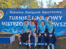 Na tle niebieskiego banneru z napisem Szkolny Związek Sportowy, Turniej Finałowy Mistrzostw Mazowsza stoi ośmiu chłopców w strojach reprezentacji szkoły.