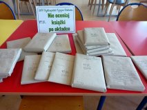 Na czerwonym stole rozłożone prostokątne przedmioty zapakowane w szary papier, w tle na kartce a4 napis: "Nie oceniaj książki po okładce".
