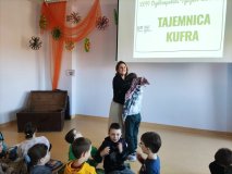 Biblioteka, w tle napis Tajemnica kufra, pod ścianą stoi stara, brązowa skrzynia, nauczycielka jednemu z uczniów zawiązuje oczy kolorową chustą.