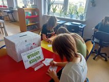 Trzy uczennice siedzą przy stoliku wyraźnie zajęte pisaniem na kartce - obok pudło z napisem Wybory Książek.