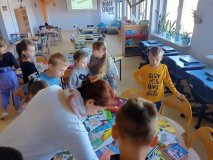 Wnętrze biblioteki, grupa uczniów stoi skupiona przy stoliku zastawionym książkami, słuchają nauczyciela.