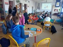pomieszczenie czytelni biblioteki, duża grupa dzieci skupiona przy kilku stolikach, grają w gry planszowe oraz budują z puzzli trasy dla małych robotów – ozobotów