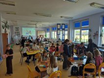 czytelnia biblioteki, duża grupa dzieci skupiona przy kilku stolikach, grają w gry planszowe oraz budują z puzzli trasy dla małych robotów – ozobotów