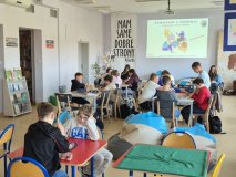 pomieszczenie czytelni biblioteki, duża grupa dzieci skupiona przy kilku stolikach, grają w gry planszowe