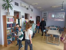 wnętrze czytelni biblioteki, kilkoro Uczniów zgromadzonych przy kole fortuny, które służy jako Tiara Przydziału, w tle na projektorze zdjęcie starej, stylowej biblioteki