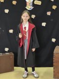Uczennica na tle czarnej ścianki z napisem „Ogólnopolski Tydzień Bibliotek”, „Hogwartowy challenge” pozuje do zdjęcia, jest przebrana za bohaterkę z serii książek o Harrym Potterze – Hermionę – czarny płaszcz z czerwonym podpiciem, czerwono-żółty krawat, różdżka w dłoni