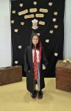 Uczennica pozuje do zdjęcia na tle czarnej ścianki z napisem „Ogólnopolski Tydzień Bibliotek”, „Hogwartowy challenge”, jest przebrana za bohaterkę z serii książek o Harrym Potterze – Hermionę – czarny płaszcz z czerwonym podpiciem, czerwono-żółty krawat, różdżka w dłoni, na głowie czarny kaptur