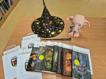 blat stołu, na którym stoi miniaturowa figurka bohatera książek o Harrym Potterze-  Zgredka oraz nagrody konkursowe – książki i zapakowane w torebki z logiem Szkoły i Rady Rodziców – karty upominkowe