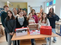 Uczniowie stoją przy ławce szkolnej z kolorowymi pudełkami walentynkowymi, ozdobionymi serduszkami i kwiatami. 