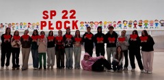 grupa młodzieży ze Szkoły Podstawowej nr 22 w Płocku stoi w szeregu w jasnej hali sportowej, trzymając czerwone papierowe serca. W tle kolorowa dekoracja z postaciami dzieci oraz duży napis 'SP 22 PŁOCK'. Uczniowie ubrani są w codzienne stroje, prezentując różnorodne style. Zdjęcie oddaje atmosferę wspólnoty i zaangażowania