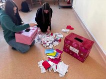 foto_1 dwie uczestniczki koła Samorządu Uczniowskiego siedzące na podłodze sali lekcyjnej, przed nimi ozdobne różowoczerwone skrzynki pocztowe z walentynkowymi motywami, z których wyjętych jest ogromna ilość kolorowych kartek walentynkowych