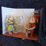 Miniaturowa scenografia do bajki „Kopciuszek”. Na tle wnętrza kamiennej chaty z kominkiem dwie figurki trollów – jeden klęczy z pantofelkiem w ręku.
