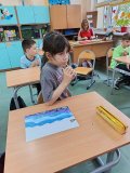 W klasie uczennica siedzi przy ławce i degustuje miód przy użyciu drewnianego patyczka. Na ławce leży kolorowa praca plastyczna utrzymana w odcieniach niebieskiego i fioletu oraz złoty piórnik. W tle widać innych uczniów siedzących przy ławkach oraz nauczycielkę pracującą przy biurku. Atmosfera sugeruje spokojne zajęcia edukacyjne związane z pszczołami i produktami pszczelimi.