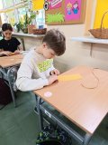 Chłopiec siedzi przy szkolnej ławce i wykonuje świecę z wosku pszczelego, podczas gdy pozostali uczniowie uczestniczą w tych samych warsztatach w klasie.