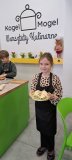 Dziewczynka stoi z talerzem, na którym znajdują się samodzielnie przygotowane przez nią warzywne wrapy. W tle widoczne jest logo pracowni „Kogel-Mogel” – miejsca warsztatów kulinarnych.