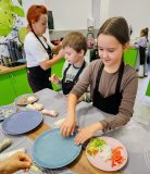 Dziewczynka składa swoje warzywne rolsy, obok niej stoi chłopiec, który przygotował już trzy rolsy, a przy nich znajduje się pani wspomagająca dzieci w trakcie warsztatów.