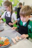 Chłopiec składa swoje warzywne rolsy; przygotował już trzy z nich. Obok niego stoi jego koleżanka. Dziewczynka również przygotowuje i składa swoje warzywne danie.