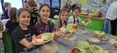 Cztery dziewczynki podnoszą talerze z przygotowanymi rozdrobnionymi warzywami oraz serem. Produkty te wykorzystają do przygotowania warzywnych wrapów.