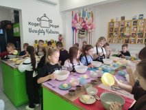 Uczniowie klasy pierwszej przygotowują się do pracy, uważnie słuchając poleceń osoby prowadzącej, która za chwilę wyjaśni im kolejne etapy wykonania ciasteczek owsianych. Przed dziećmi znajdują się miski oraz wszystkie produkty potrzebne do przygotowania ciasta.