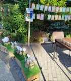 Stoisko artystyczne. Na stole, w pudełkach, znajdują się koraliki do robienia biżuterii. Nad stolikiem, na sznurkach, wiszą obrazki do kolorowania. Obok stolika, po jego lewej stronie, stoją drewniane skrzynki, a na nich, w papierowych doniczkach, umieszczone są biało-czarne kwiaty.