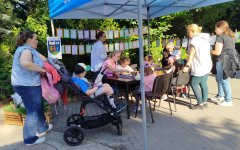 Przy stole siedzą Dzieci, które wykonują prace plastyczne. Dookoła nich są Panie, które się  im przyglądają. W pobliżu stołu, na wózku, siedzi Chłopiec, który razem z Mamą podziwia pomysłowość pracujących.
